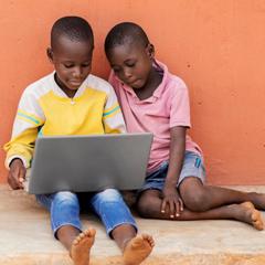 Two African boys sitting in front of an orange wall with a laptop computer